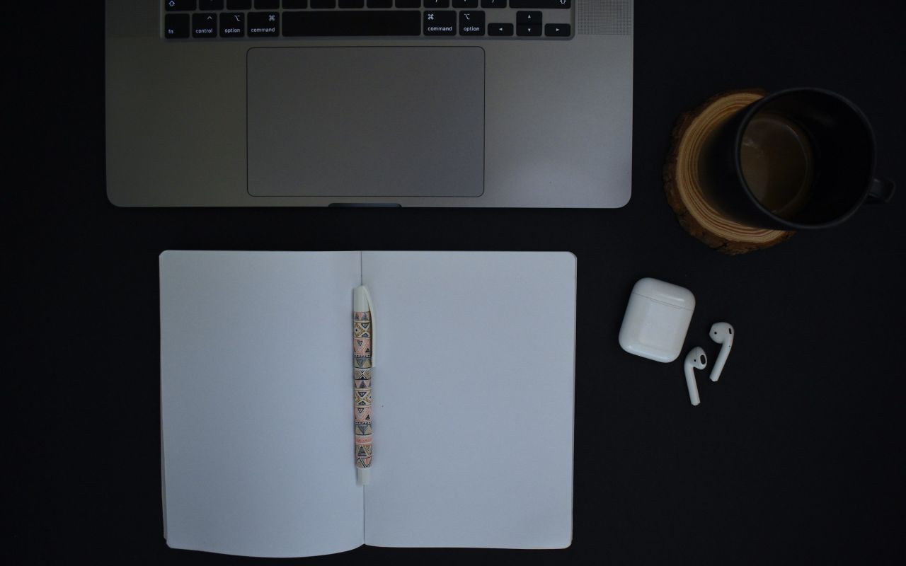 A laptop, an open notebook, a pen and a coffee mug on a dark desk